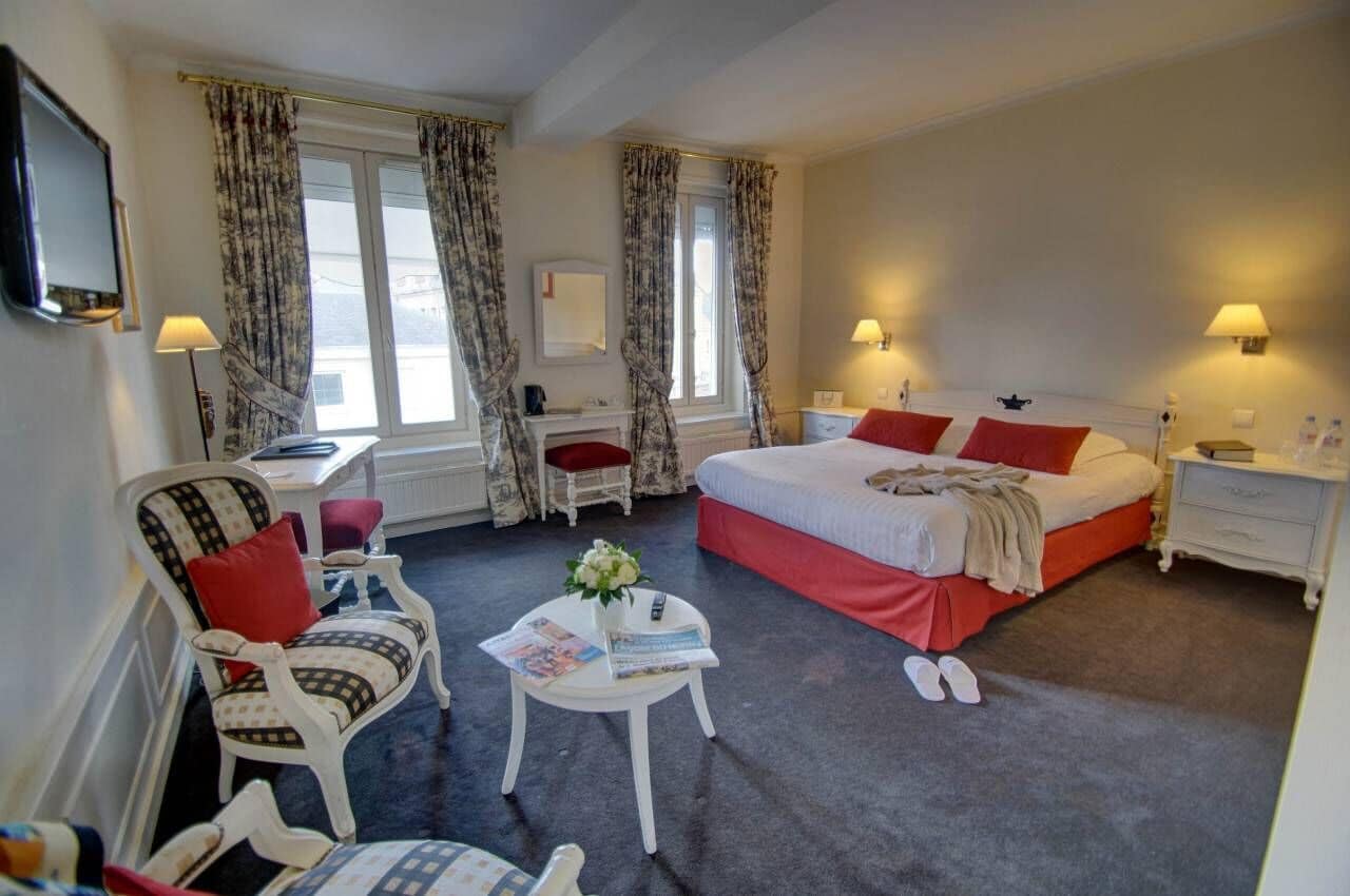 A hotel room with a bed, two chairs, a small table with a vase, and a TV on the wall.