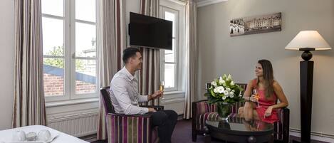 A hotel room with a large window, striped curtains, a flat-screen TV, a table with a vase of flowers, and a lamp.