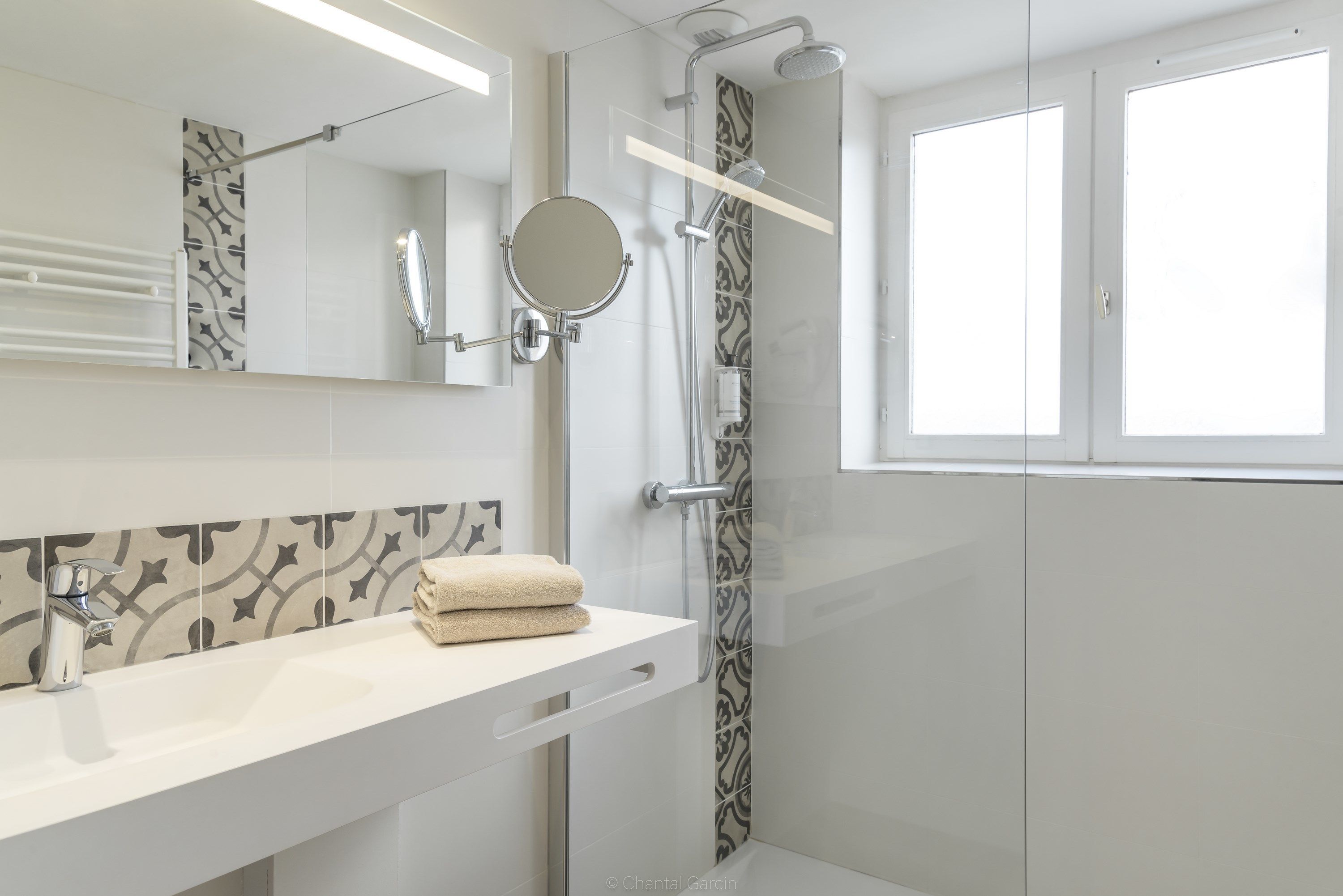 A modern bathroom with a white vanity, a round mirror, a shower with a glass door, and a patterned tile backsplash.