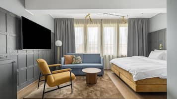 A modern hotel room with a bed, sofa, armchair, and a flat-screen TV.