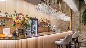 Zona de bar con mostrador de madera, taburetes, una variedad de botellas y vasos, y una estantería colgante para copas.