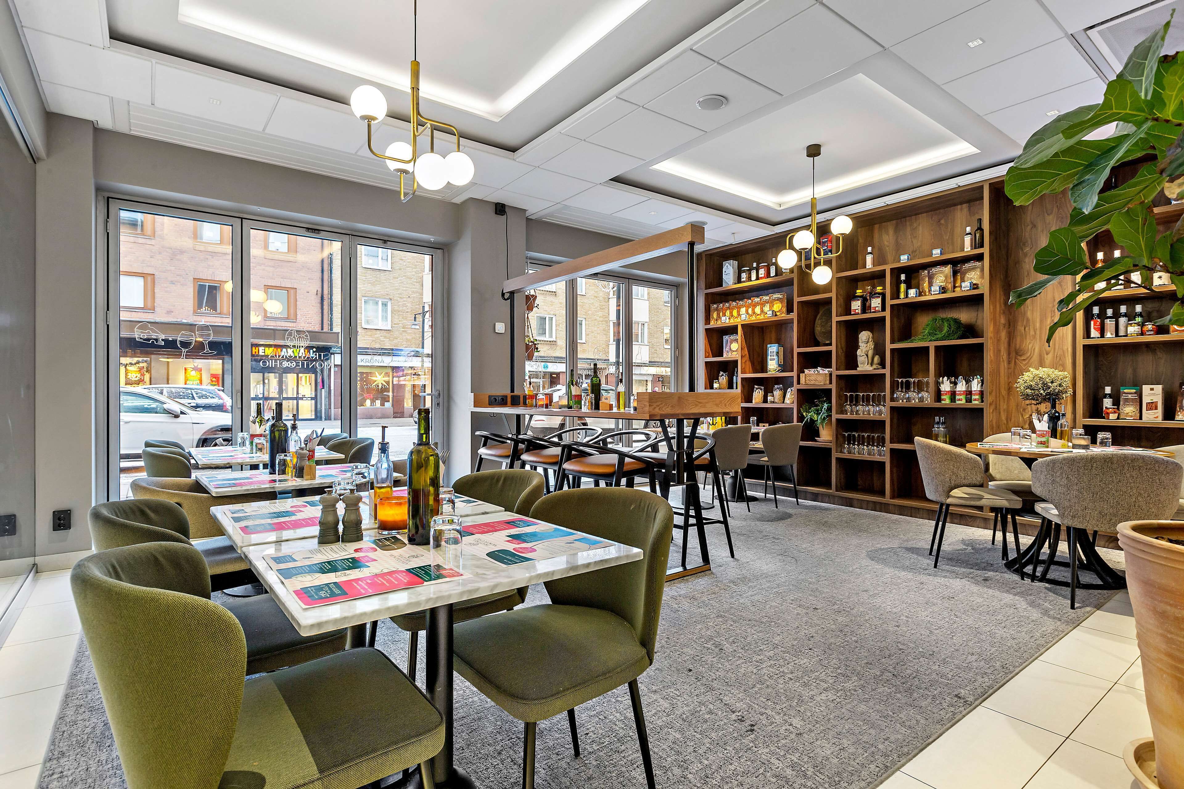 A modern restaurant interior with a central dining table, green upholstered chairs, and a wooden bookshelf with various items.