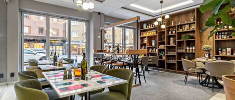Ein modernes Restaurant-Interieur mit einem zentralen Esstisch, grünen gepolsterten Stühlen und einer Holzbücherregal mit verschiedenen Gegenständen.