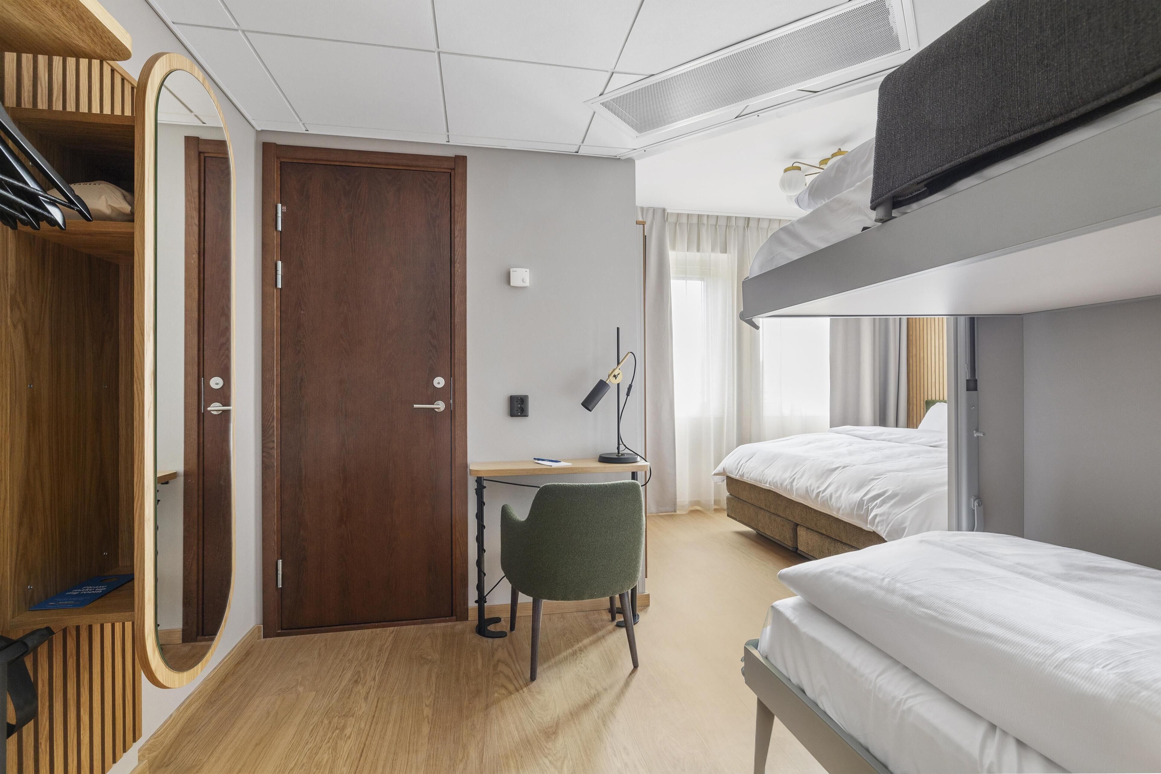 A hotel room with a bunk bed, a desk with a chair, a wardrobe, and a mirror.