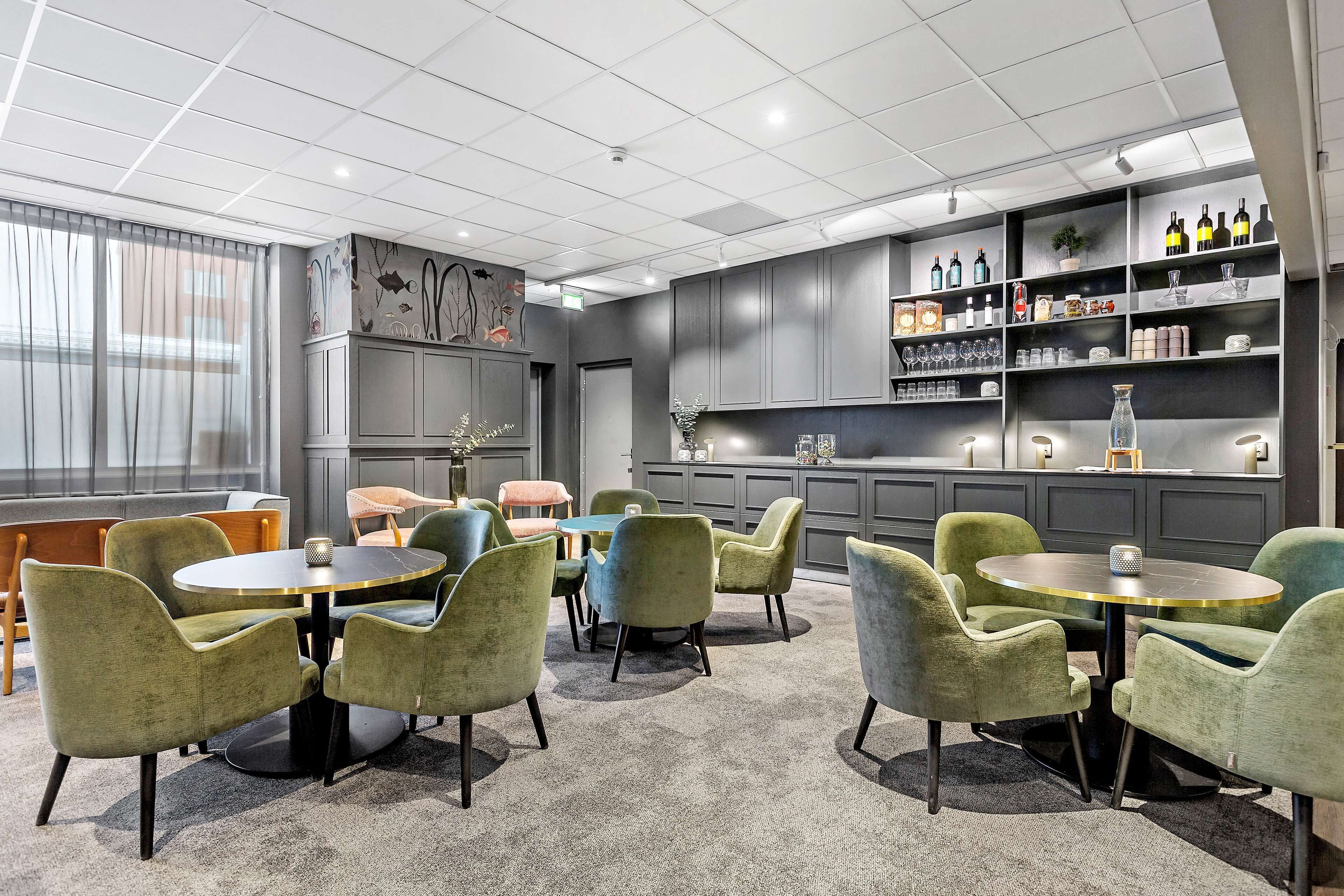 A modern lounge area with round tables and green upholstered chairs, a bar area with shelves of bottles, and a decorative mural on the wall.