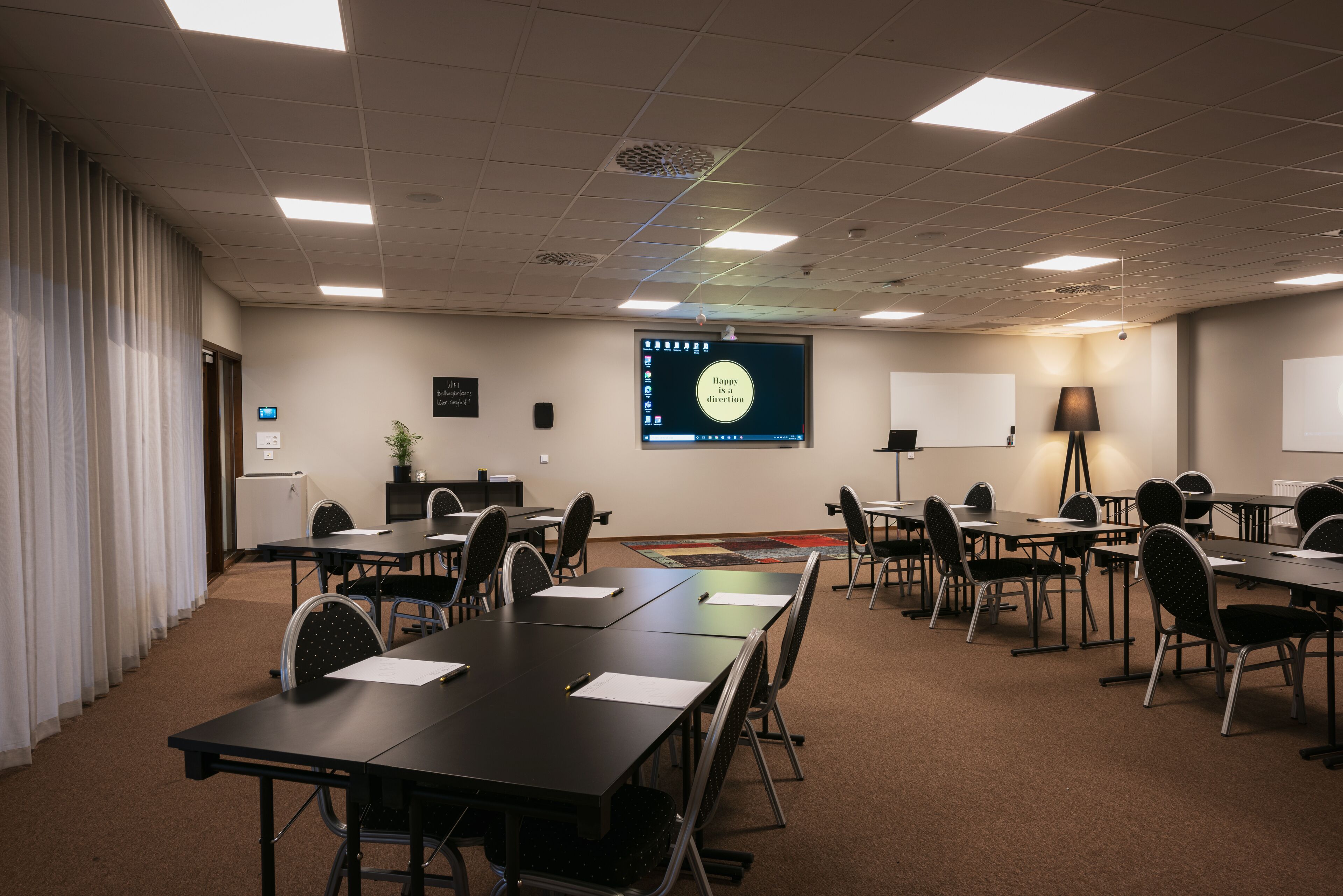 A conference room with a large screen displaying a presentation, multiple tables and chairs, a projector, and a whiteboard.
