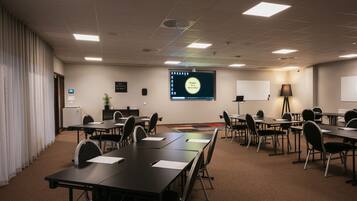 A conference room with a large screen displaying a presentation, multiple tables and chairs, a projector, and a whiteboard.