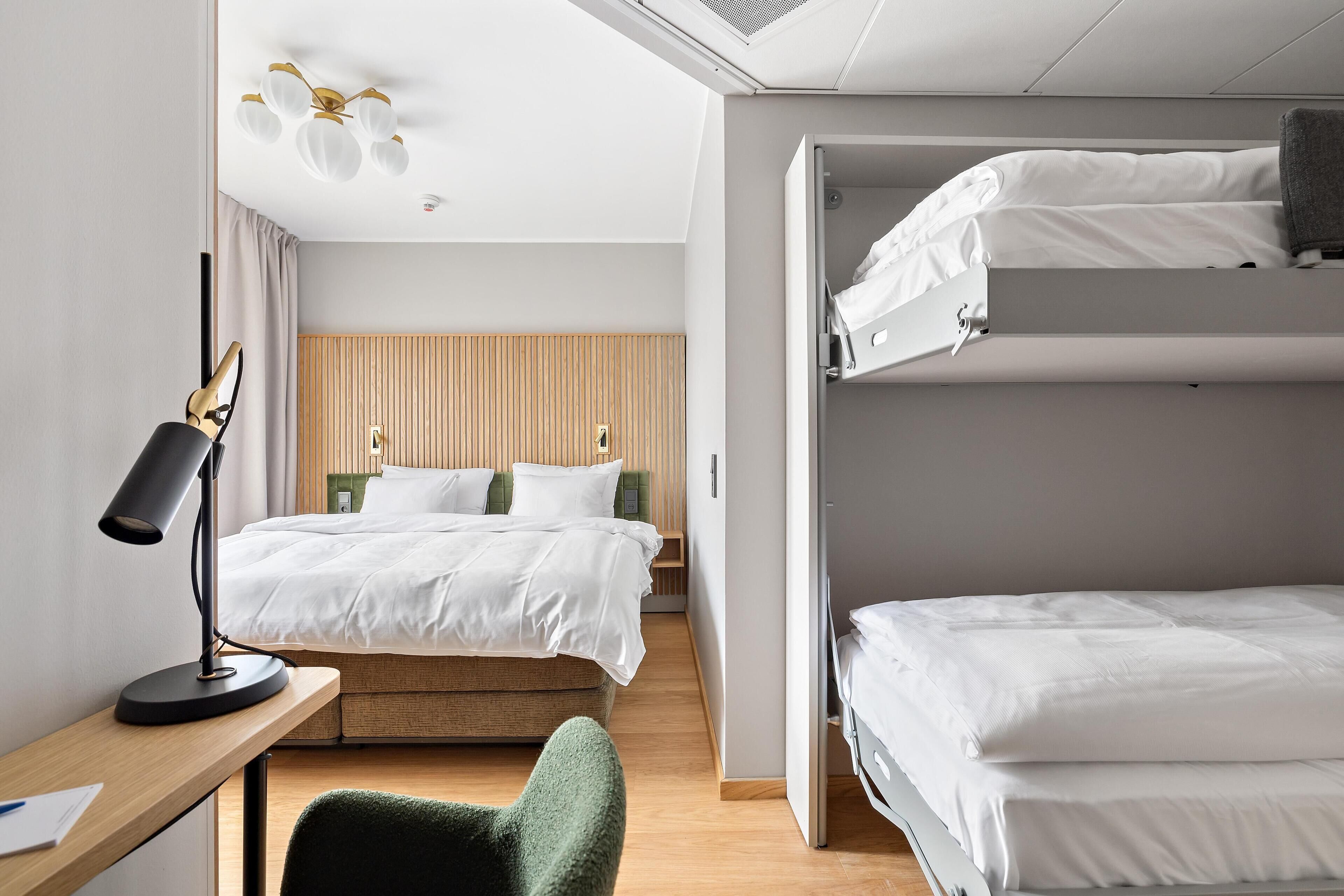 A hotel room with a bunk bed, a desk with a lamp, a chair, and a wooden headboard.