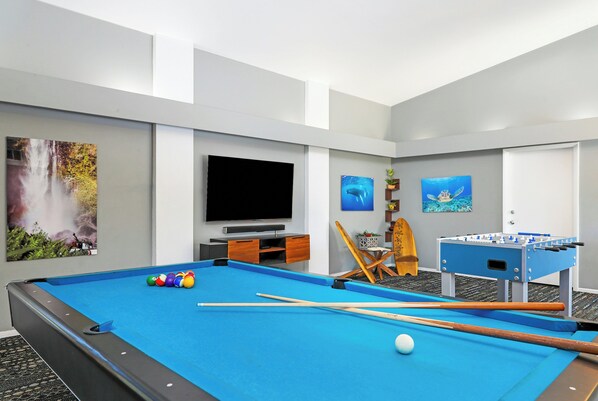 A pool table with balls, a foosball table, and a television in a room with a waterfall painting and a turtle painting on the wall. - Paniolo Greens At Waikoloa (Waikoloa)