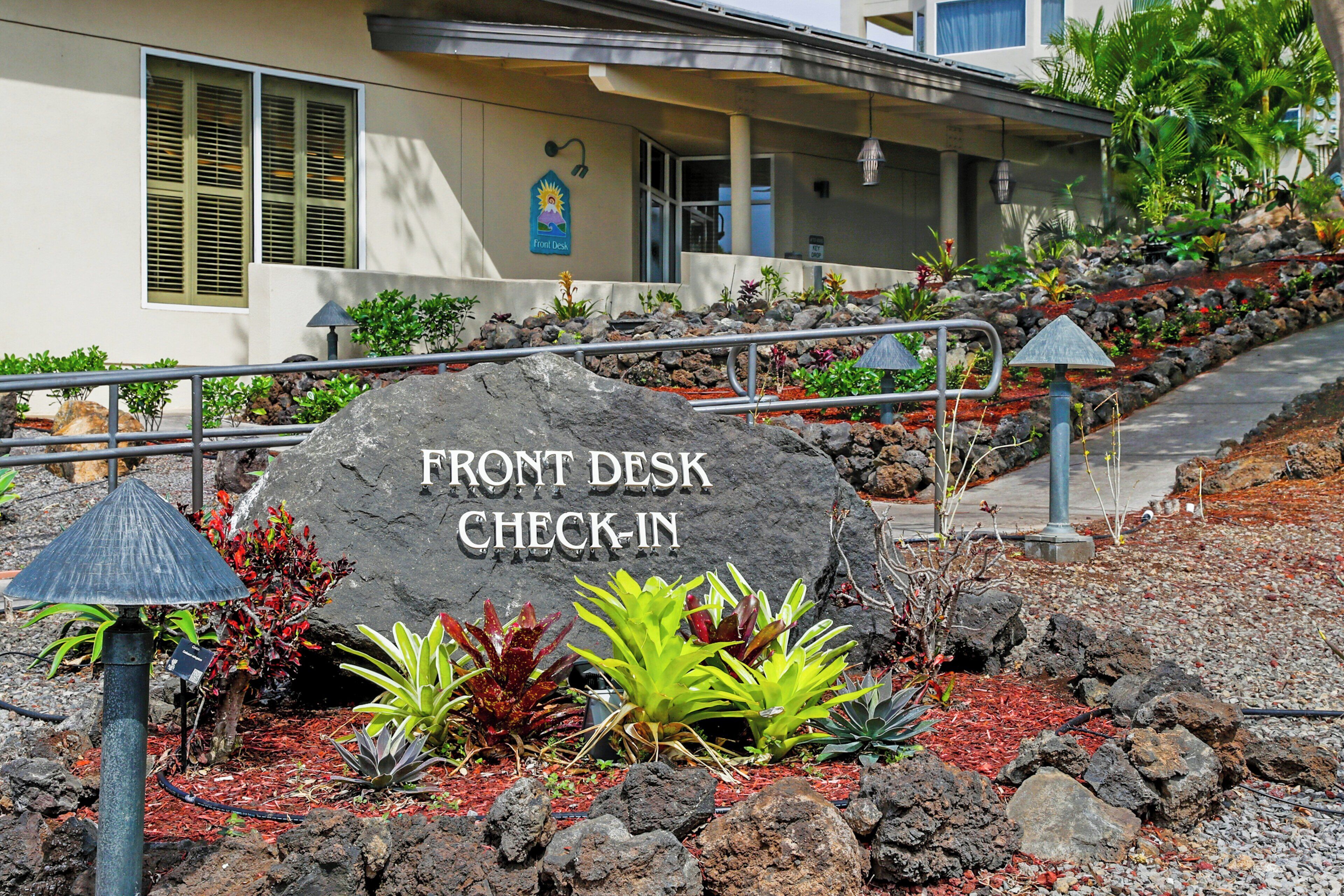 A hotel entrance with a large rock sign reading 'Front Desk Check-In,' surrounded by tropical plants and a metal railing.