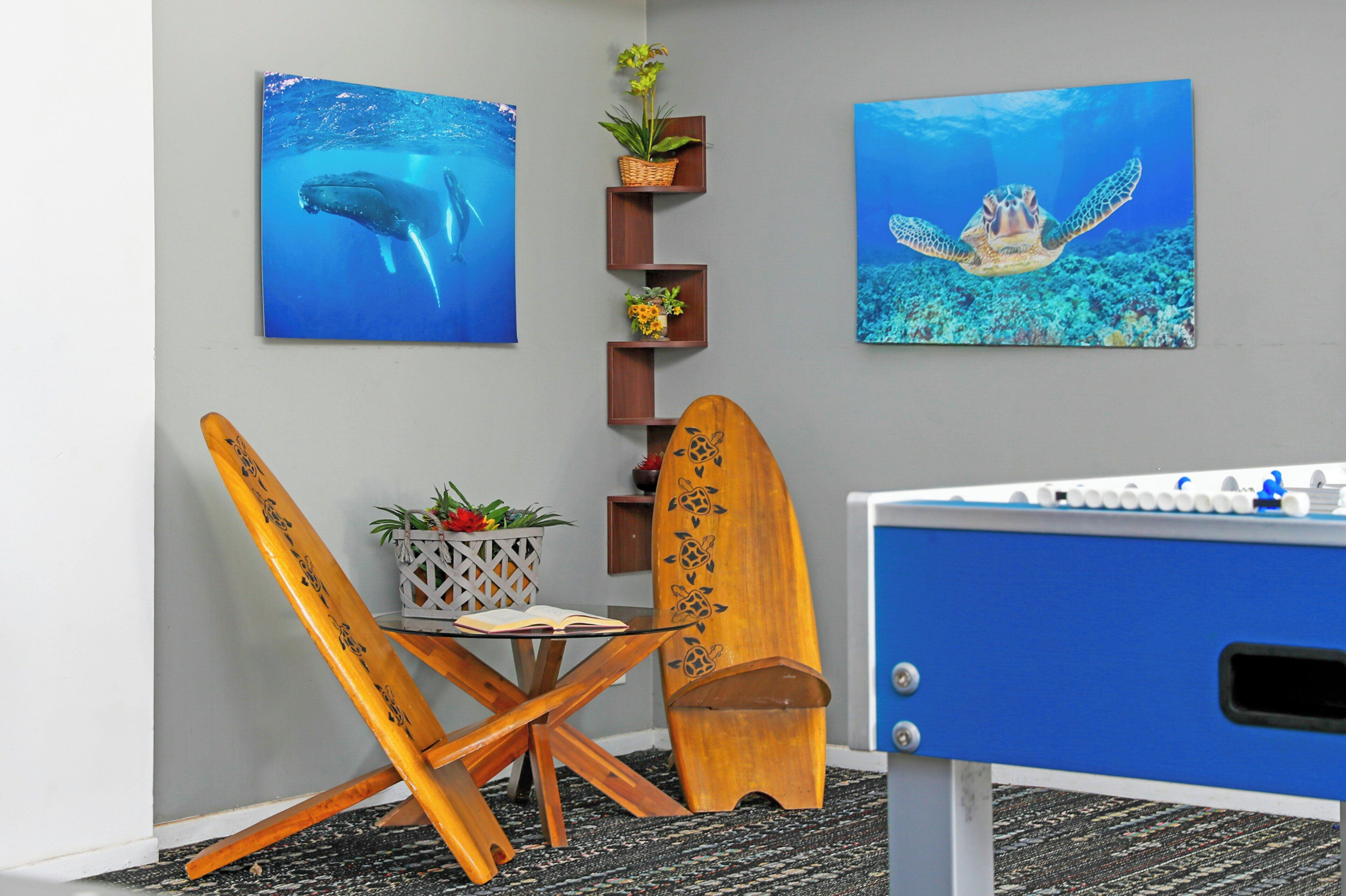 Une pièce comprenant une table de baby-foot, deux planches de surf en bois, une petite table avec un livre et une plante, ainsi que deux tableaux encadrés représentant des animaux marins, accrochés au mur.