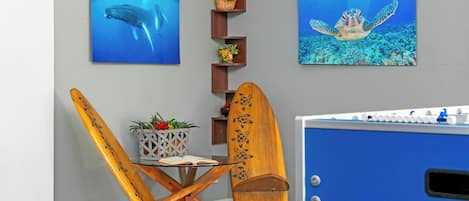Una habitación con una mesa de futbolín, dos tablas de surf de madera, una mesita con un libro y una planta, y dos cuadros enmarcados con motivos marinos en la pared.