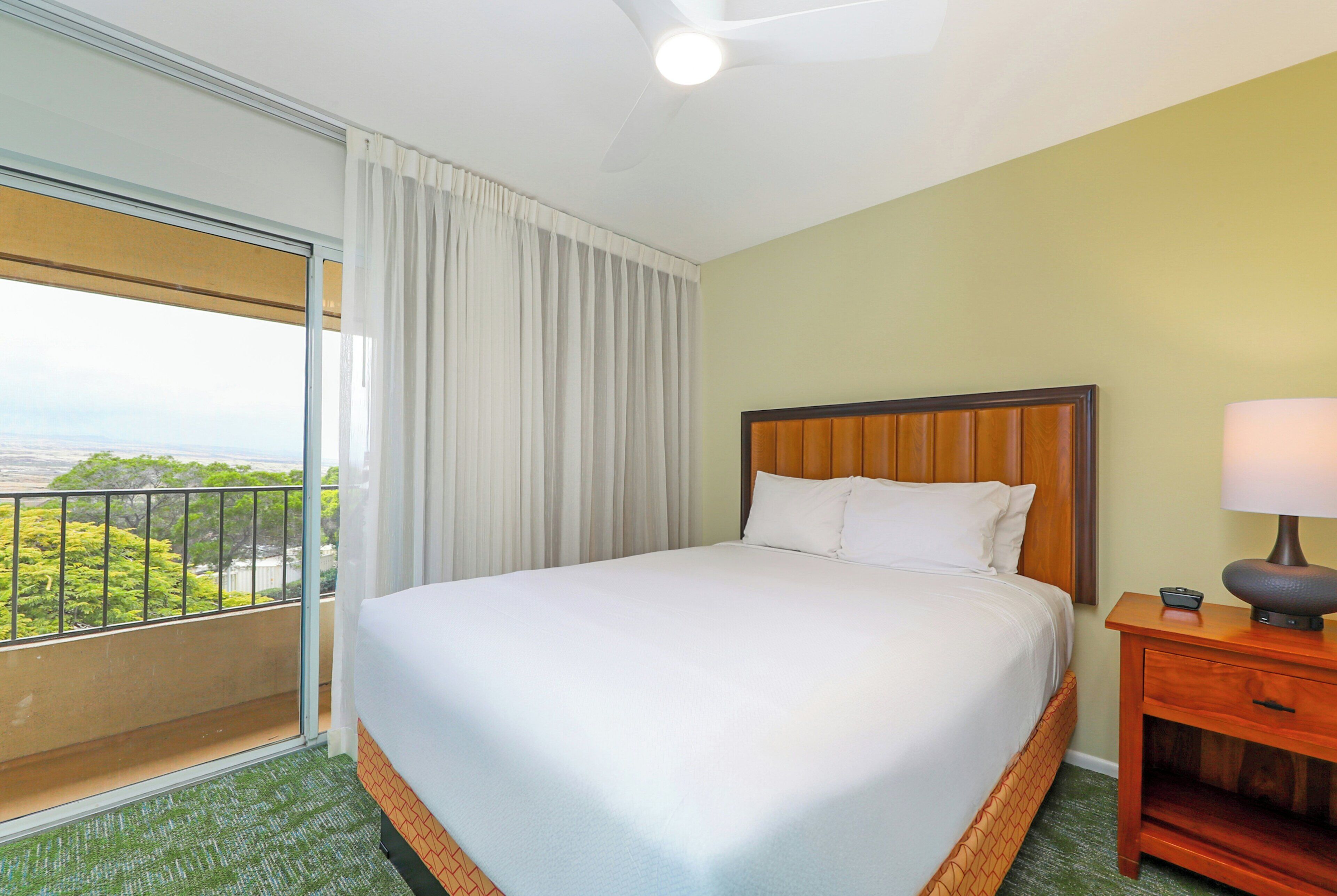 Habitación de hotel con una cama grande, cabecera de madera, mesita de noche con lámpara y vistas a vegetación por la ventana.