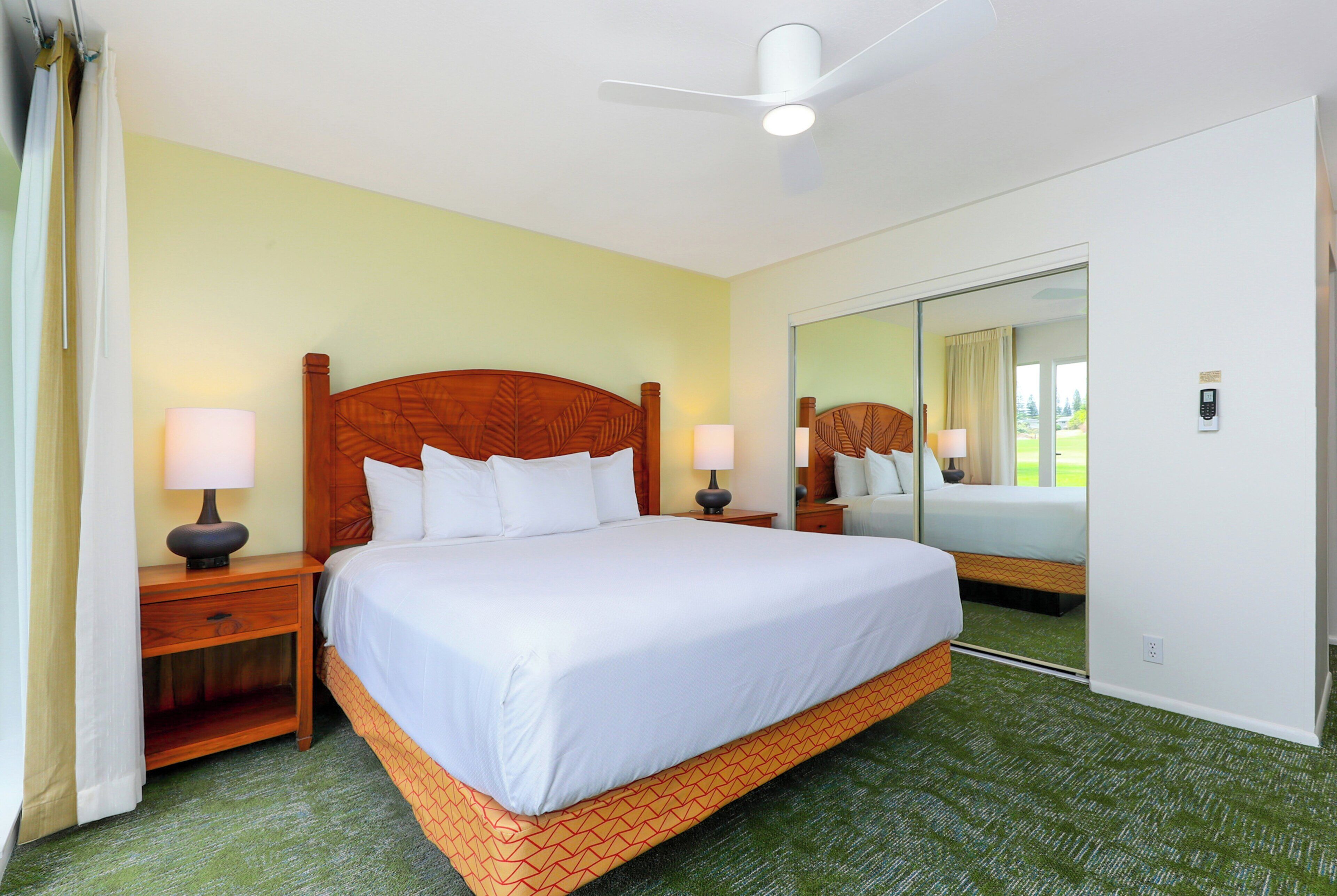 A hotel room with a large bed, two bedside tables with lamps, a ceiling fan, a mirror, and a view of the outdoors through a window.