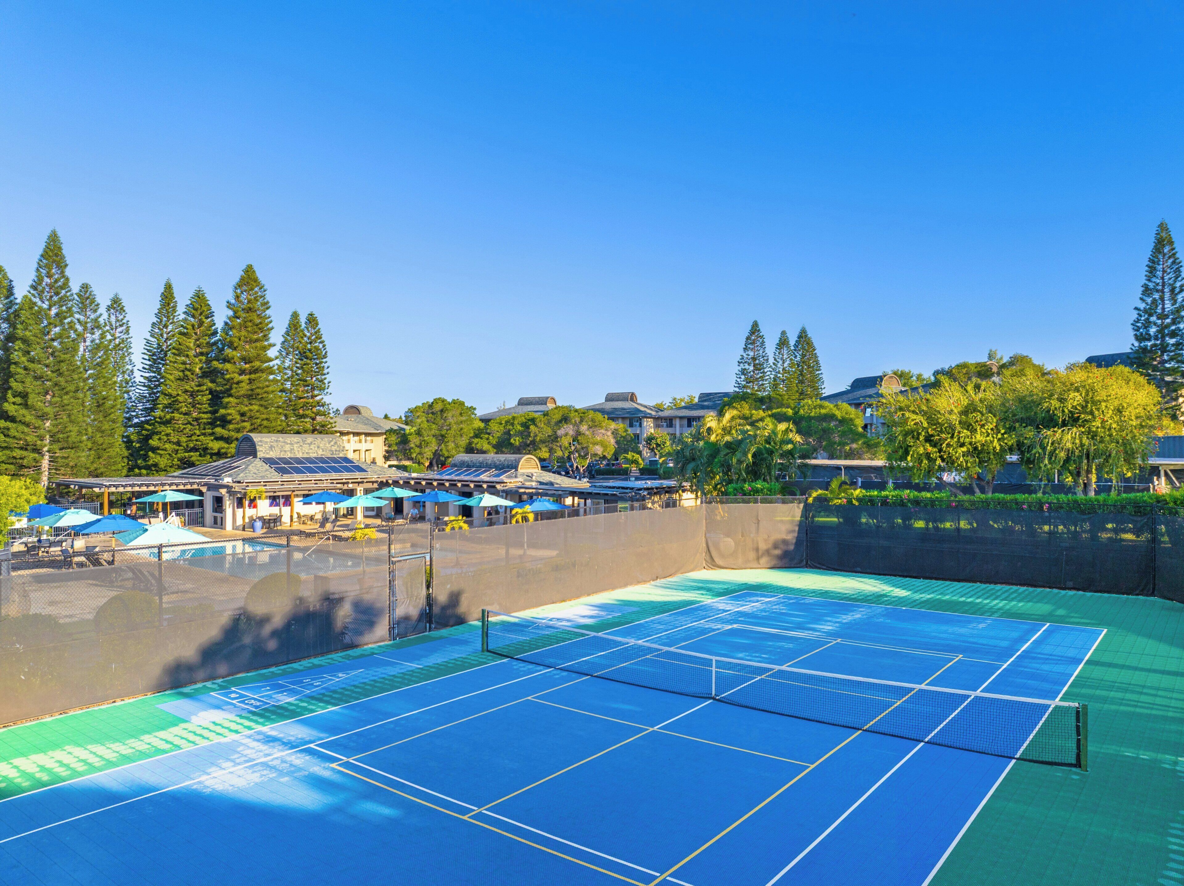 Un court de tennis en plein air, doté d’un filet et entouré d’une clôture, avec un bâtiment à toit en pente en arrière-plan.