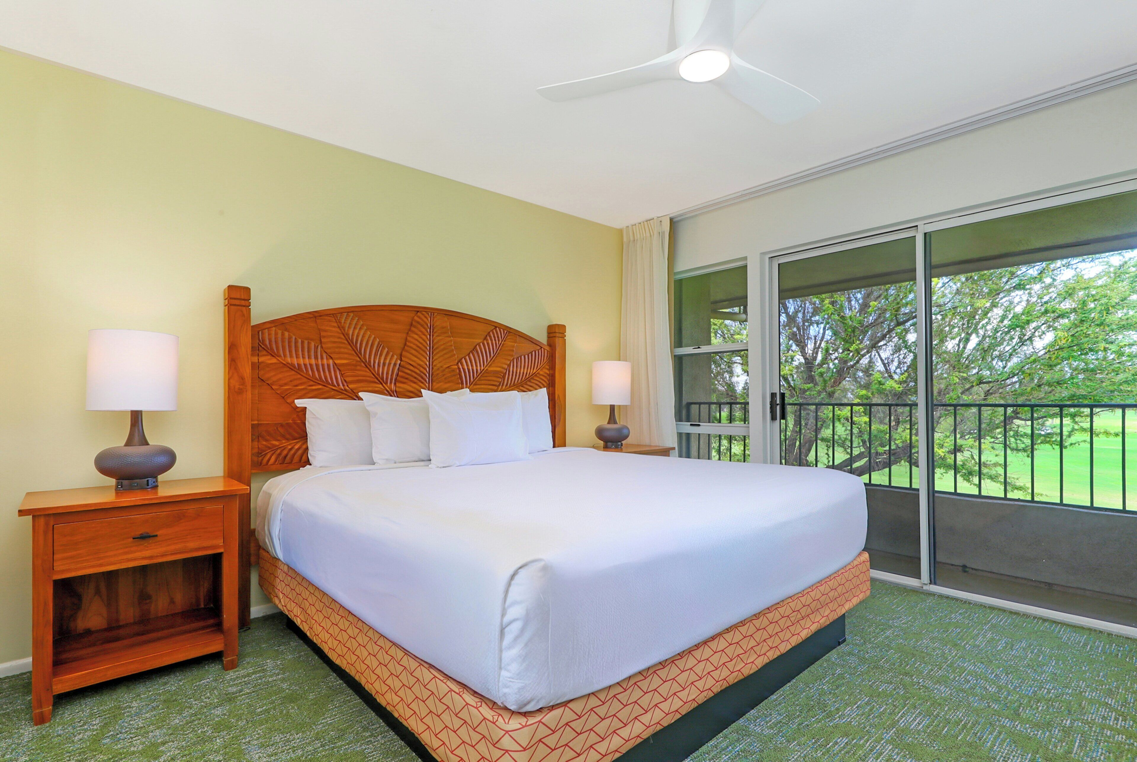 A hotel room with a large bed, two bedside tables, a ceiling fan, and a balcony with a view of trees.