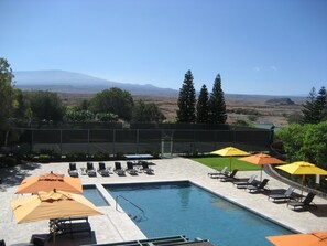 A pool area with lounge chairs and umbrellas, surrounded by a fence, with a view of mountains in the distance. - Paniolo Greens At Waikoloa (Waikoloa)