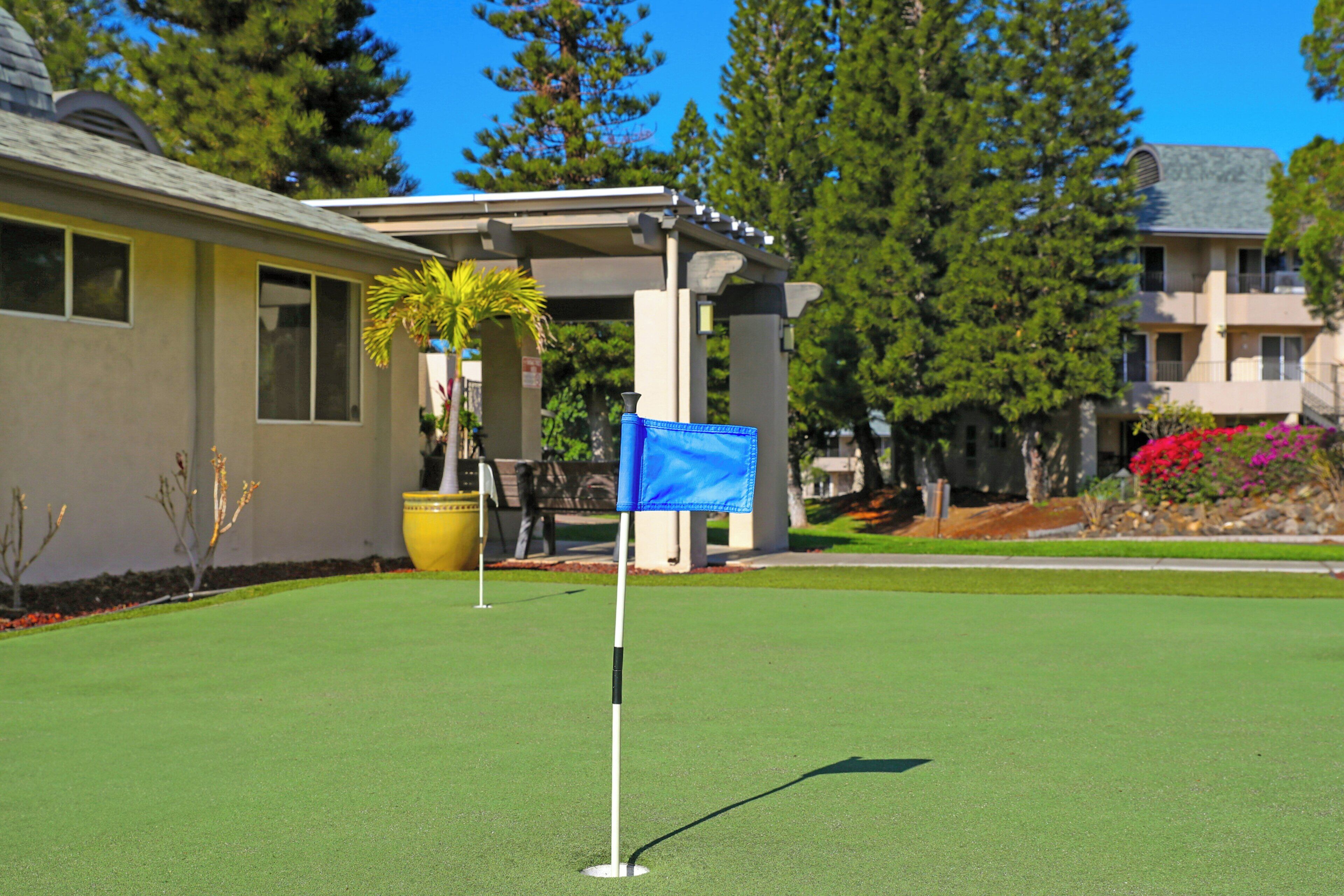 A golf course with a flagstick, a building with a porch, and a large tree.