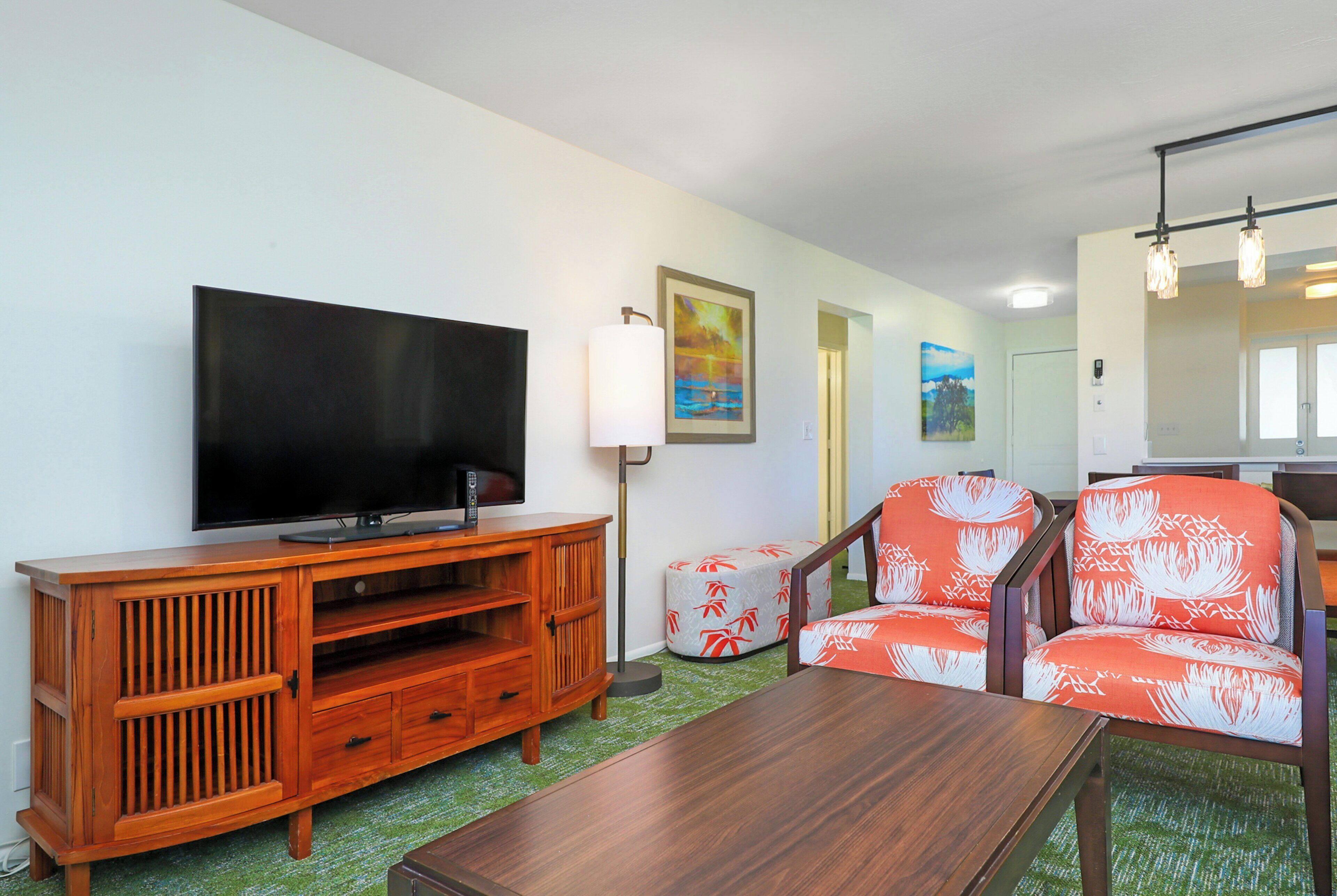 A living room with a flat-screen TV, wooden entertainment unit, two patterned armchairs, a coffee table, and a painting on the wall.
