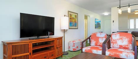 A living room with a flat-screen TV, wooden entertainment unit, two patterned armchairs, a coffee table, and a painting on the wall.