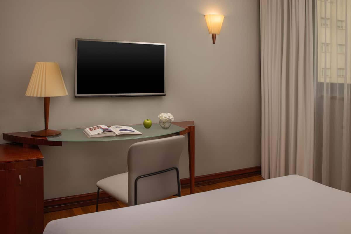 A hotel room with a bed, a desk with a lamp, a TV, and a window with curtains.