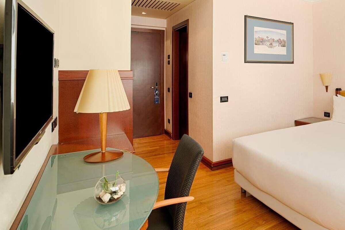 A hotel room with a bed, a desk with a lamp, a TV, and a picture on the wall.