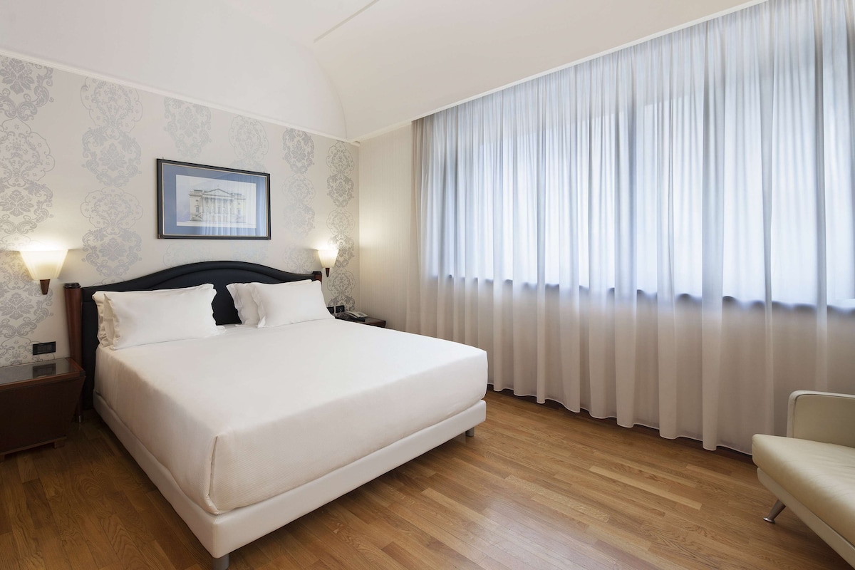 Une chambre d’hôtel avec un grand lit, deux lampes de chevet, une table de chevet, une chaise et une fenêtre avec des rideaux légers.