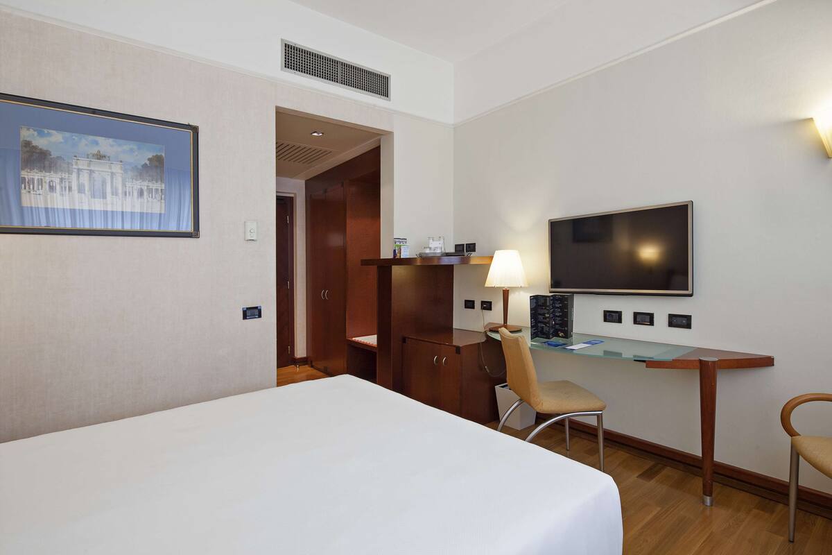 A hotel room with a bed, a desk with a lamp, a TV, and a chair.