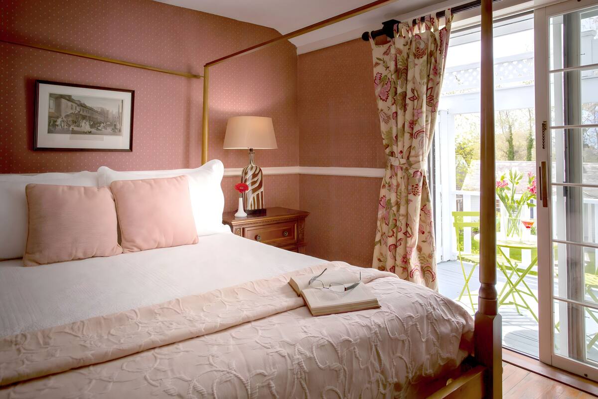 A neatly made bed with a white comforter and pink pillows, a bedside table with a lamp, and a window with floral curtains.