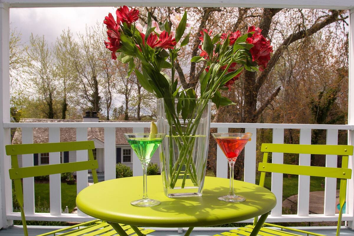 A table with a vase of red flowers, two martini glasses, and a lemon wedge on a green table, surrounded by yellow chairs on a white deck with a white railing.