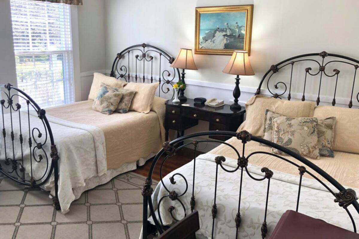 A room with two beds, each with a metal frame and a beige bedspread. There is a painting on the wall above the beds and a window with blinds on the left side.