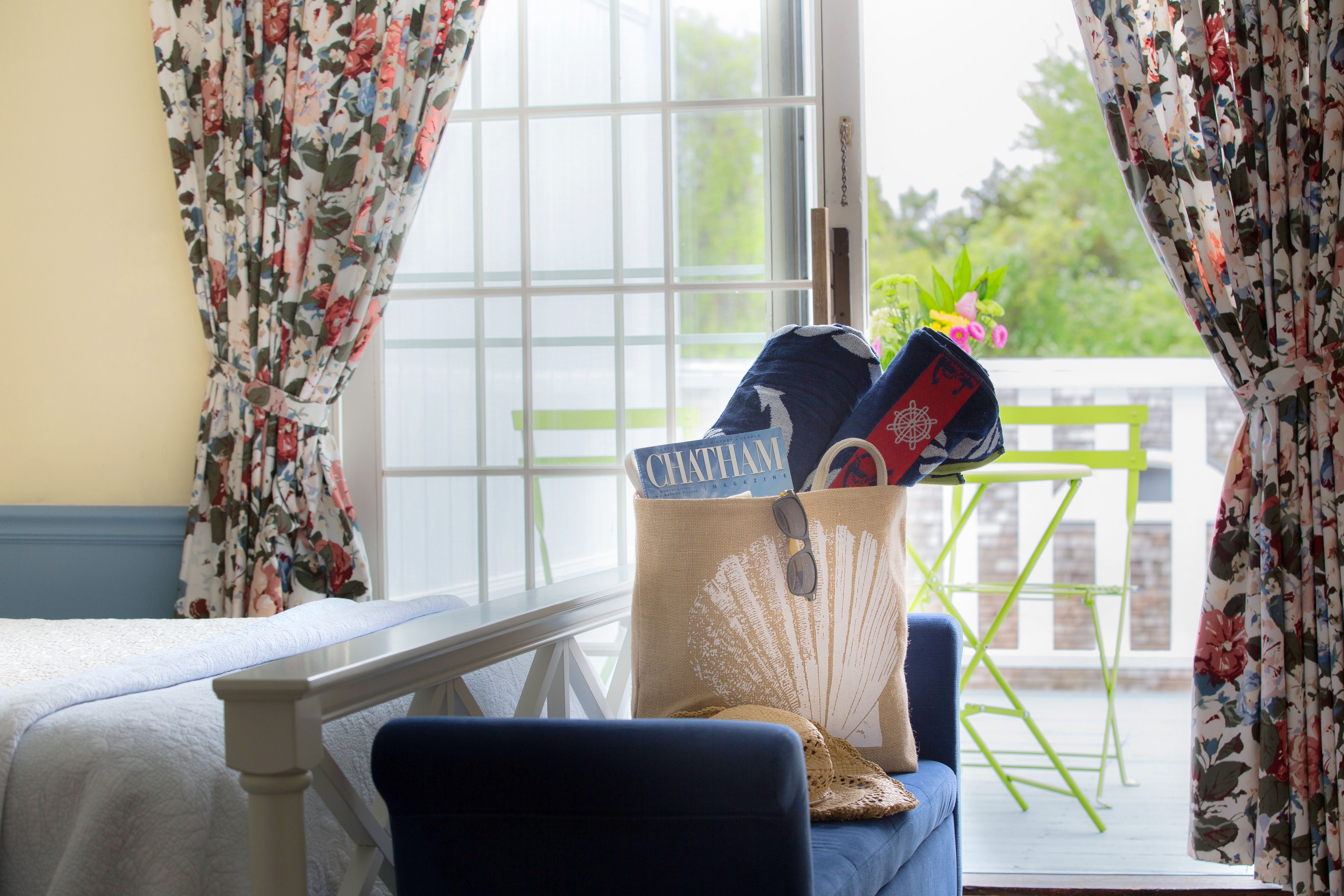 A room with a bed, a chair, a window with floral curtains, and a table with a bag and a book.