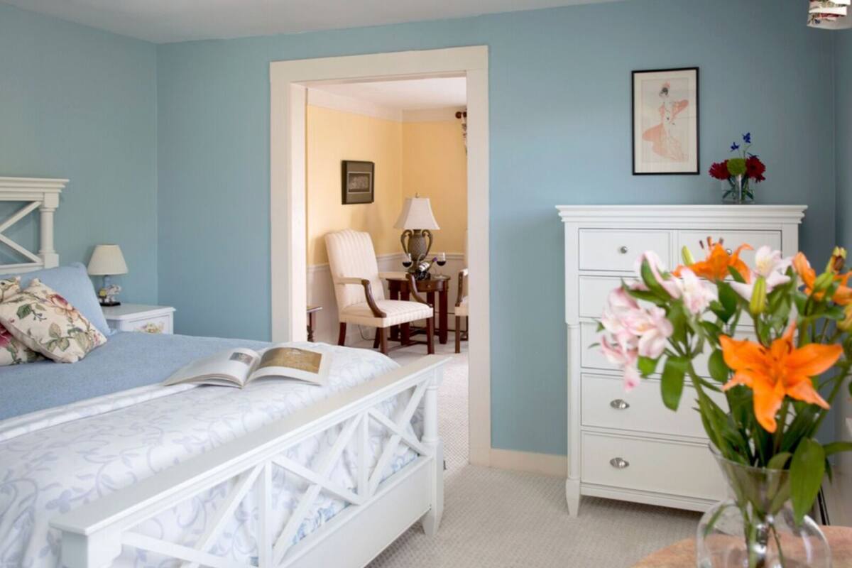 A bedroom with a bed, a dresser, a chair, and a table with a vase of flowers.