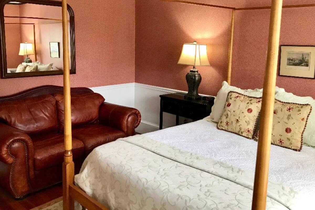 A bedroom with a four-poster bed, a brown leather sofa, a nightstand with a lamp, and a framed picture on the wall.