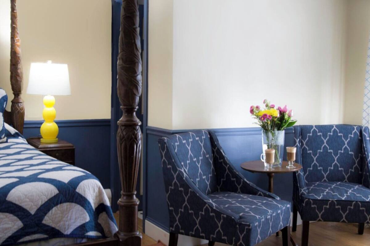 A bedroom with a four-poster bed, a nightstand with a lamp, two patterned armchairs, and a small table with a vase of flowers and two glasses of a beverage.