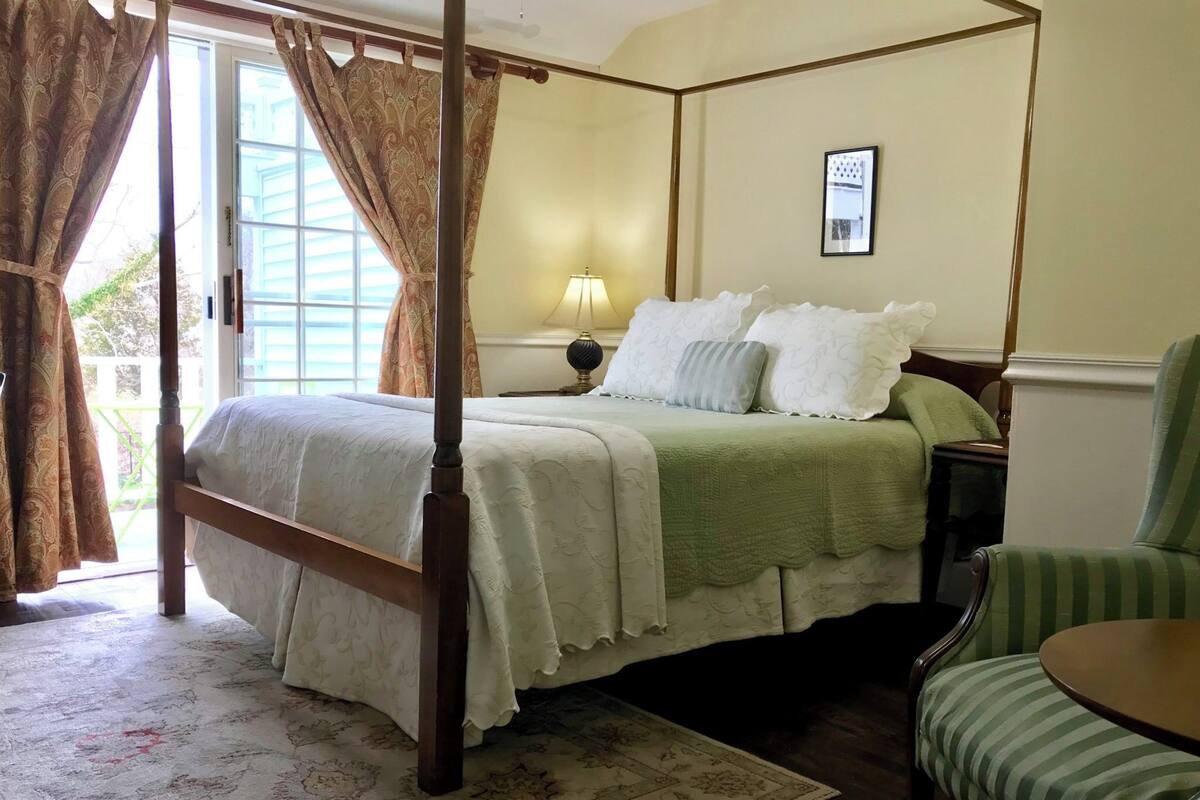 A four-poster bed with a green bedspread, a bedside table with a lamp, and a chair with a striped cushion.