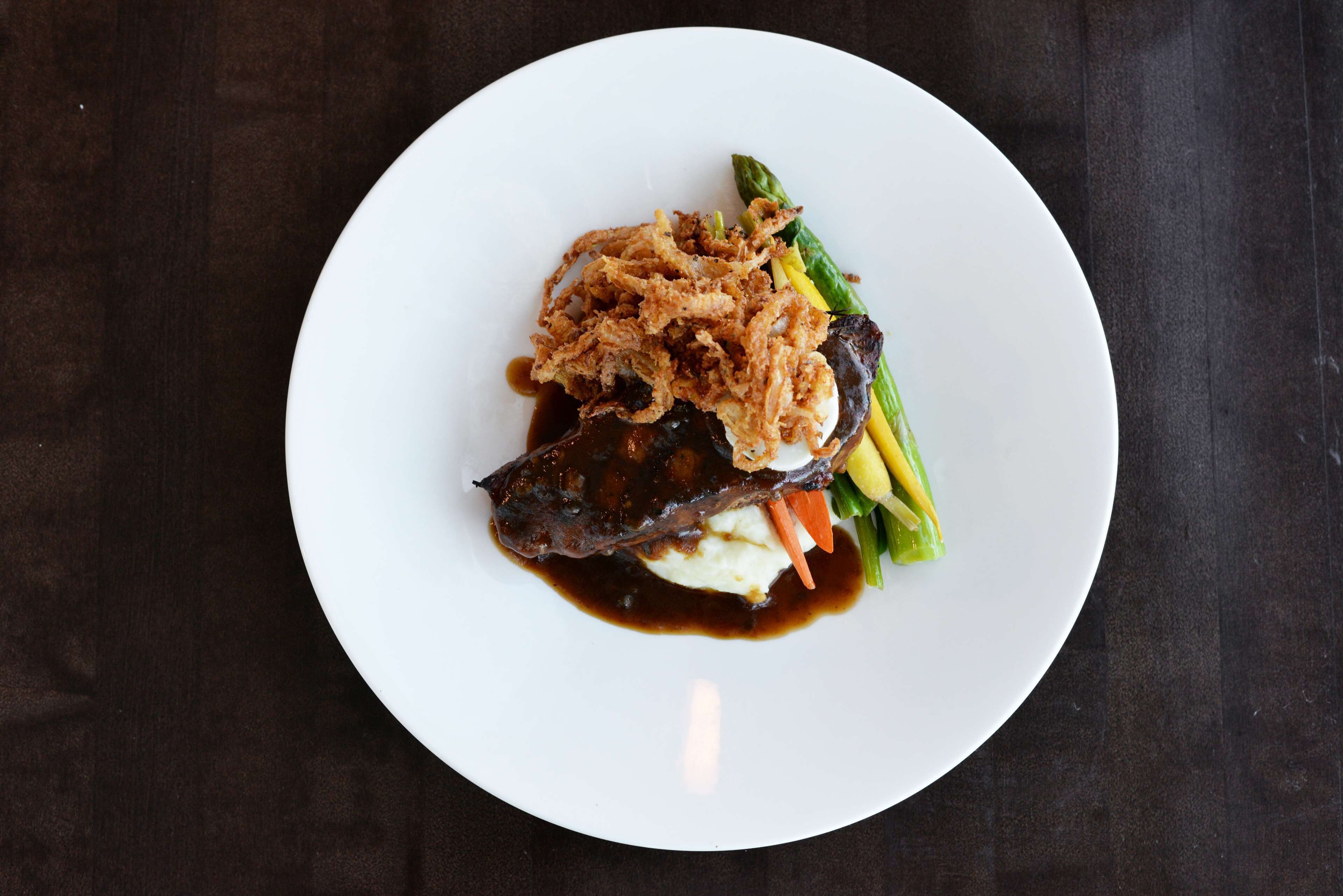 A white plate with a piece of meat, mashed potatoes, and vegetables on a dark surface.