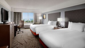 A hotel room with two beds, a desk, a TV, and a large window with a city view.