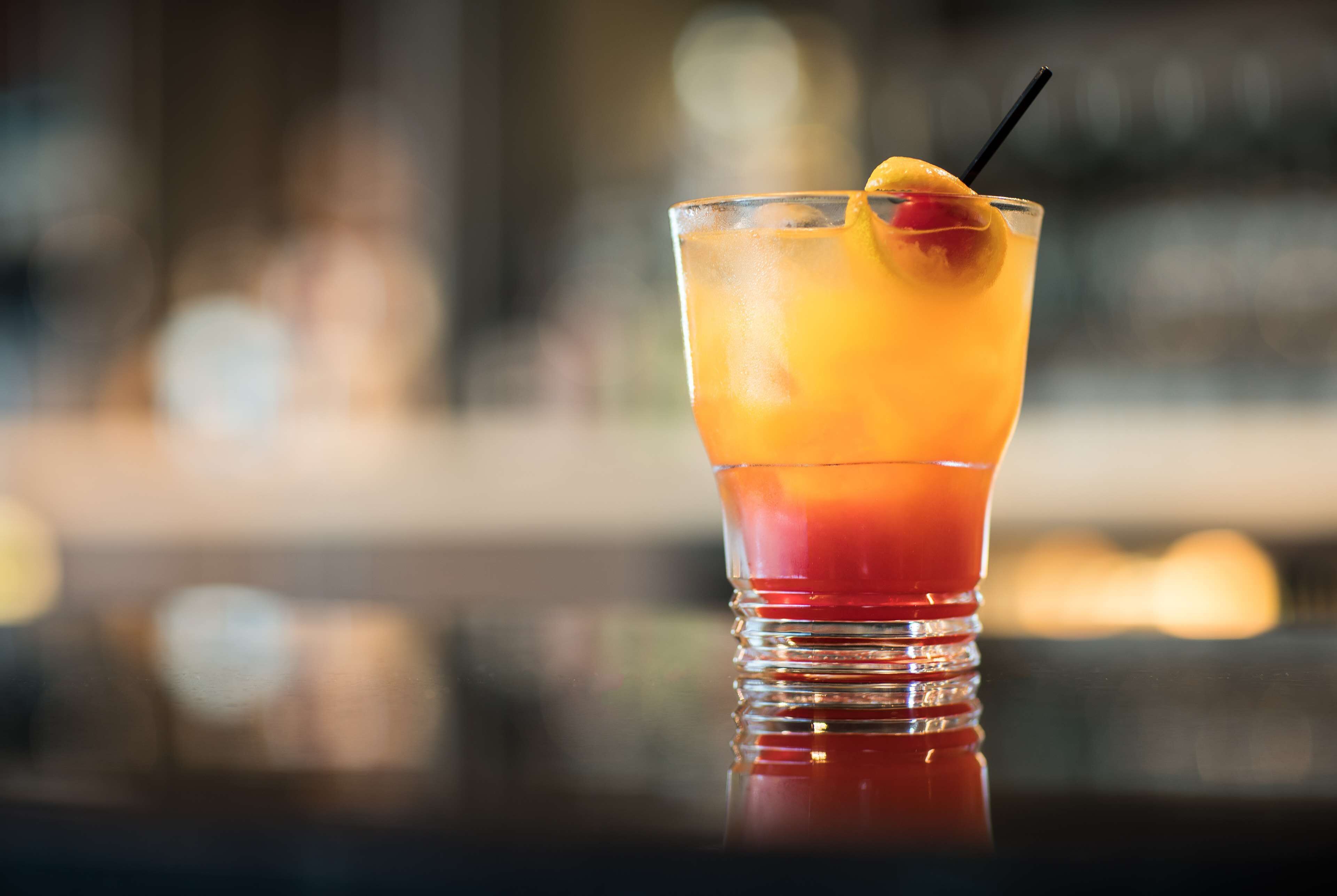 A glass of layered cocktail with a cherry garnish on a reflective surface.