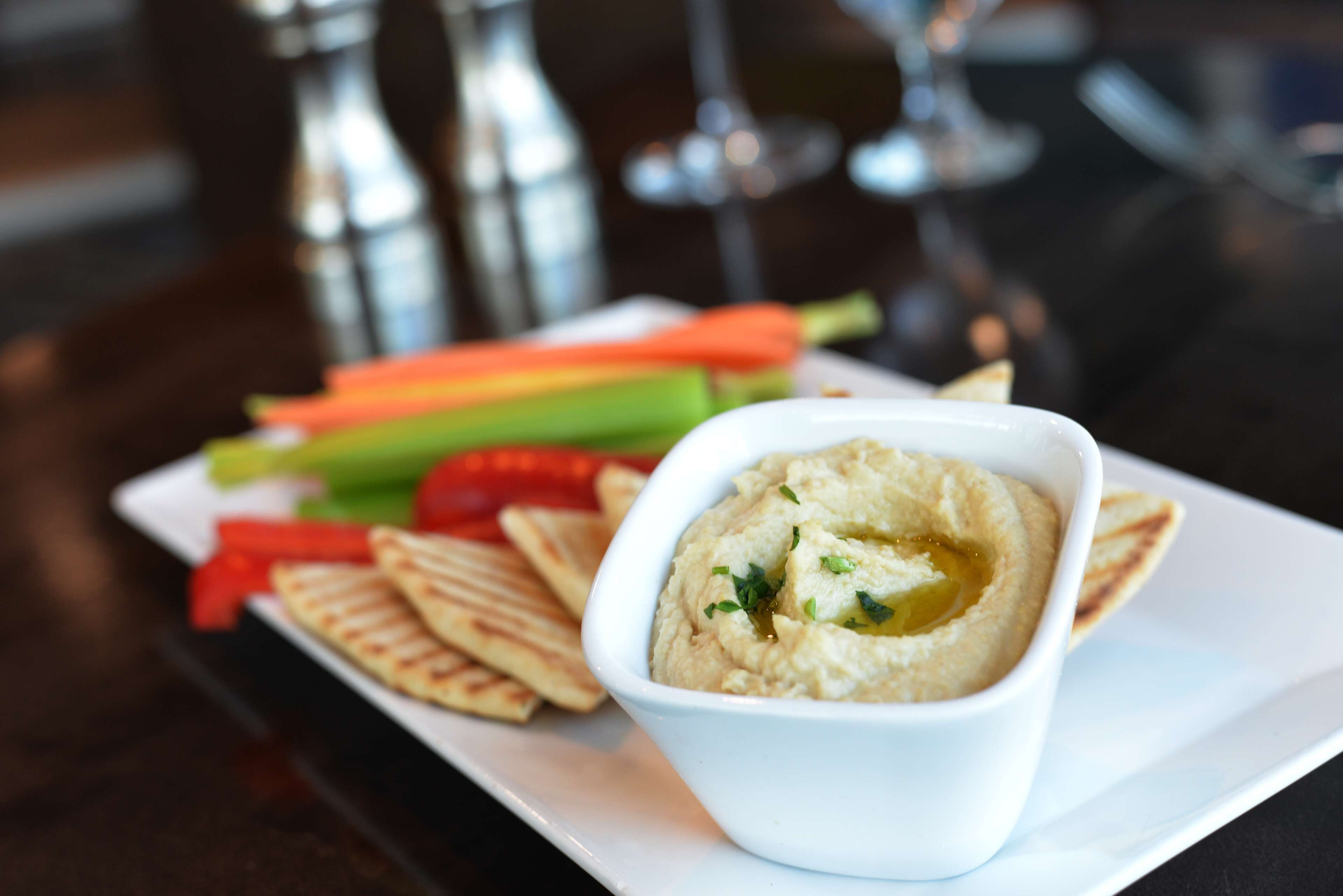 Un bol blanc de houmous, arrosé d’huile d’olive et parsemé d’herbes, servi avec du pain pita, des carottes, du céleri et des poivrons rouges sur une assiette blanche.