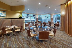 Een ruime eetzaal van een hotel met meerdere ronde tafels en stoelen, grote ramen met uitzicht op palmbomen, en een vloerbedekking.