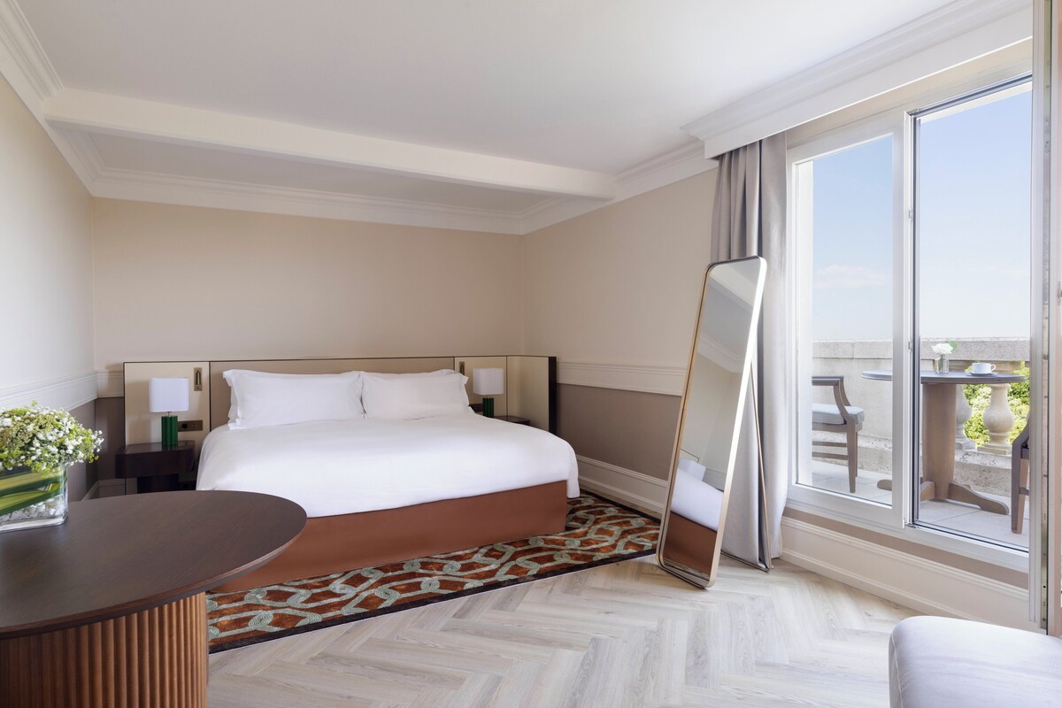 Una habitación de hotel con una cama grande, una mesita de noche con un jarrón de flores, un espejo amplio y vistas al exterior a través de una puerta corrediza de vidrio.