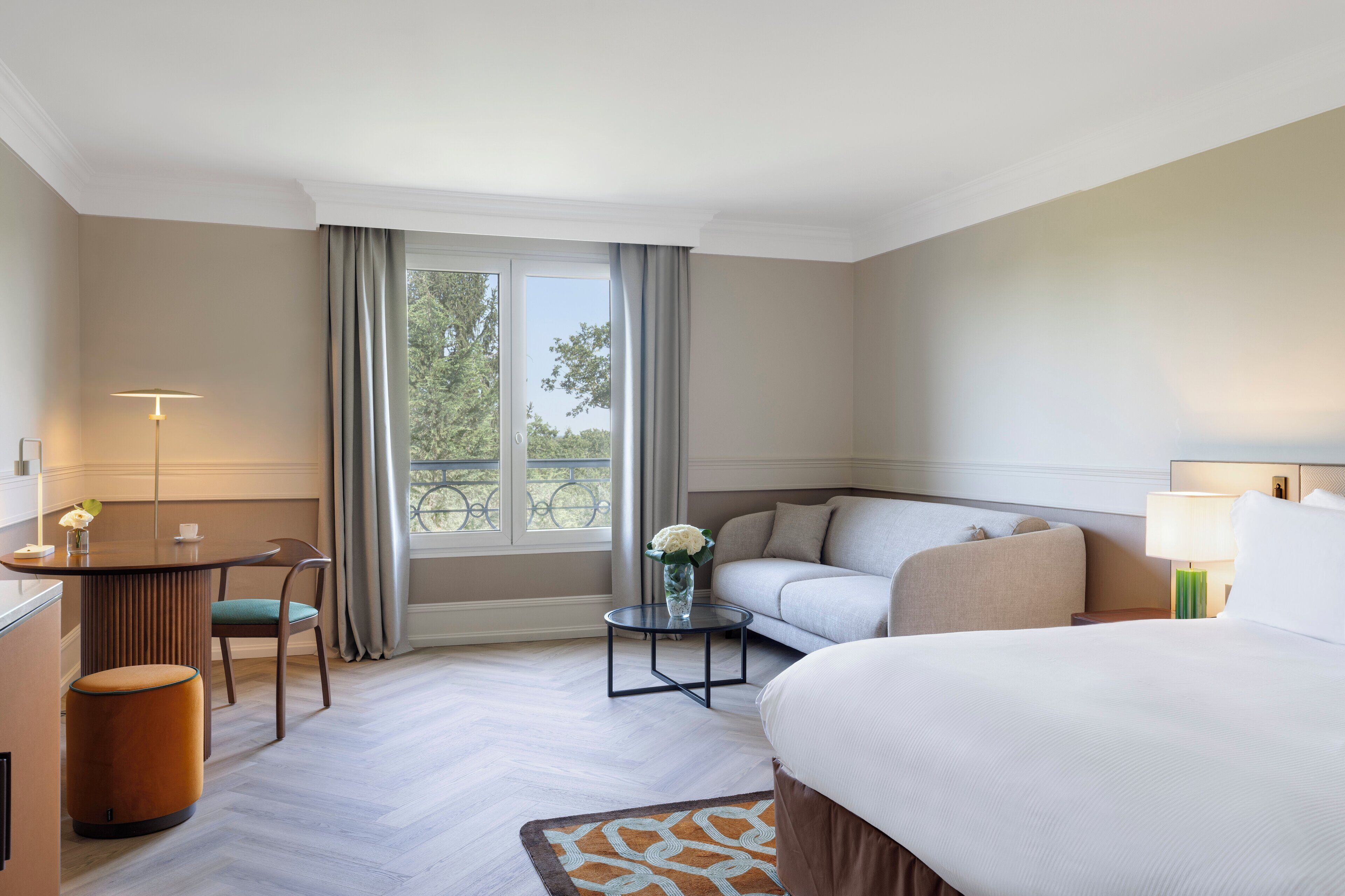 A hotel room with a bed, a sofa, a small table with a vase, a lamp, and a view of trees through the window.