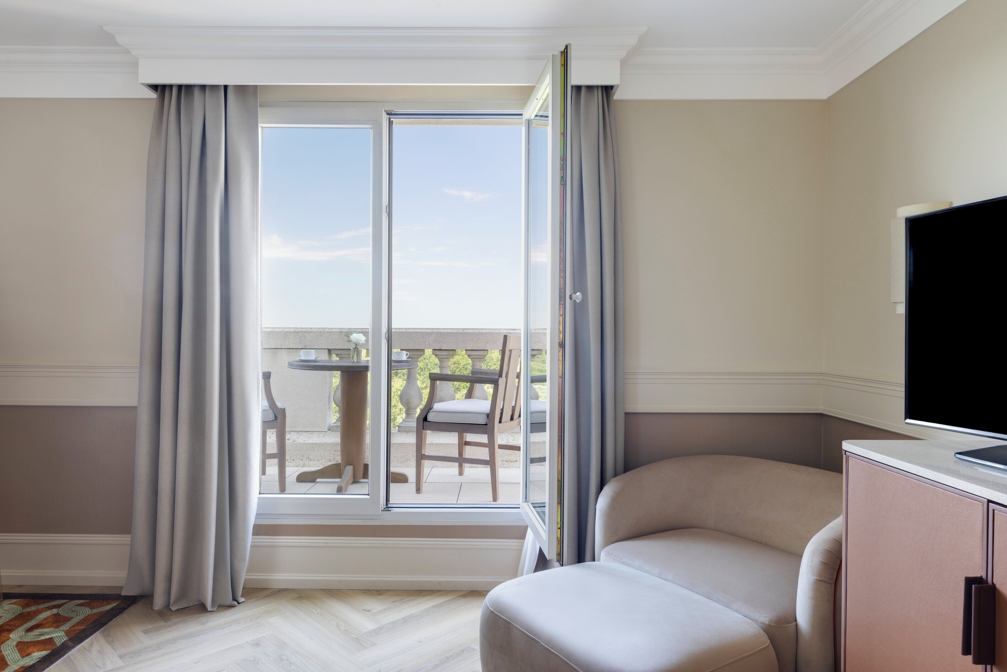 Habitación de hotel con un gran ventanal corredero que da a un balcón, un área de descanso con cojines y un televisor de pantalla plana sobre un mueble.