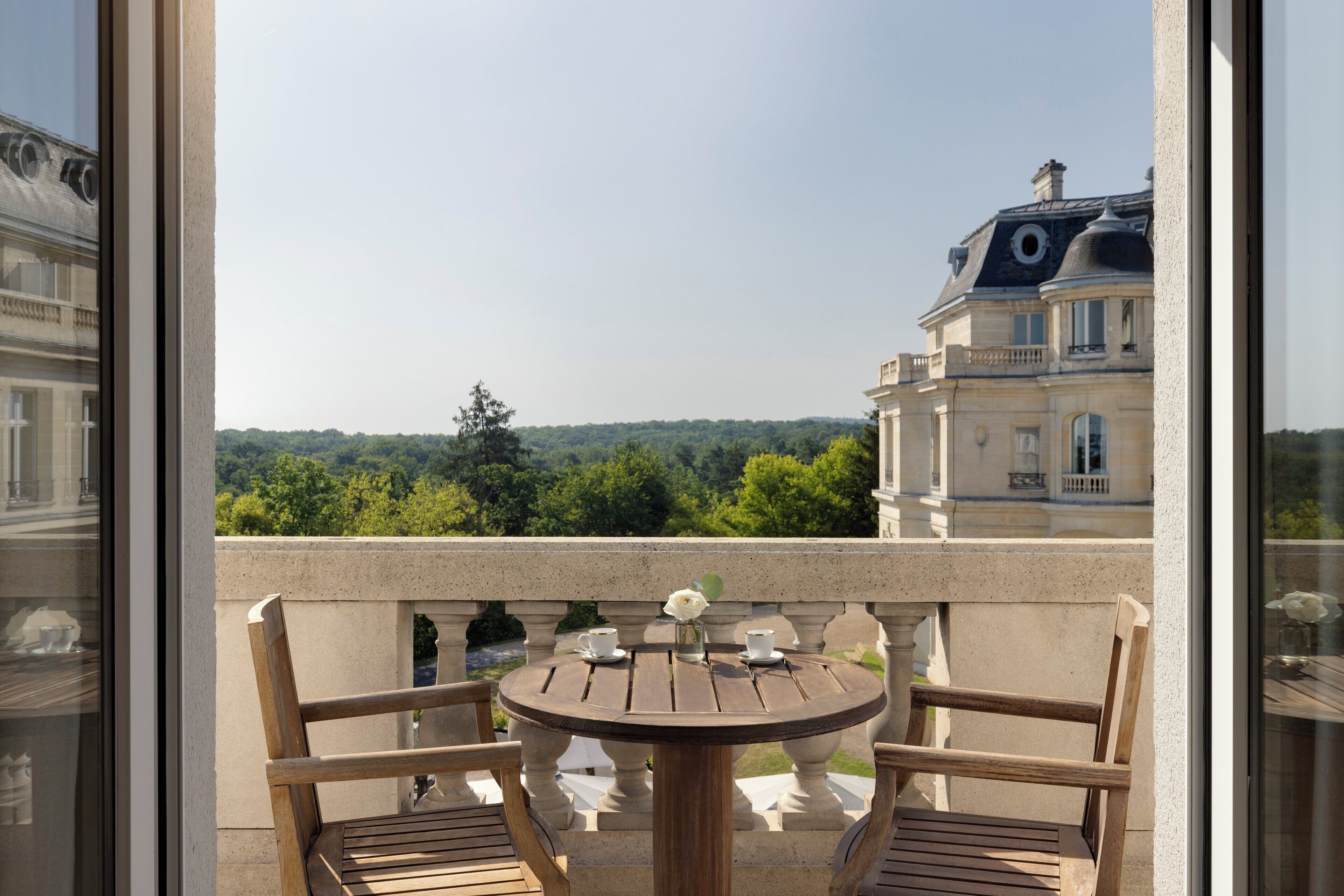 Un balcon avec une table ronde en bois et deux chaises, d’où l’on peut admirer un paysage urbain et la verdure.