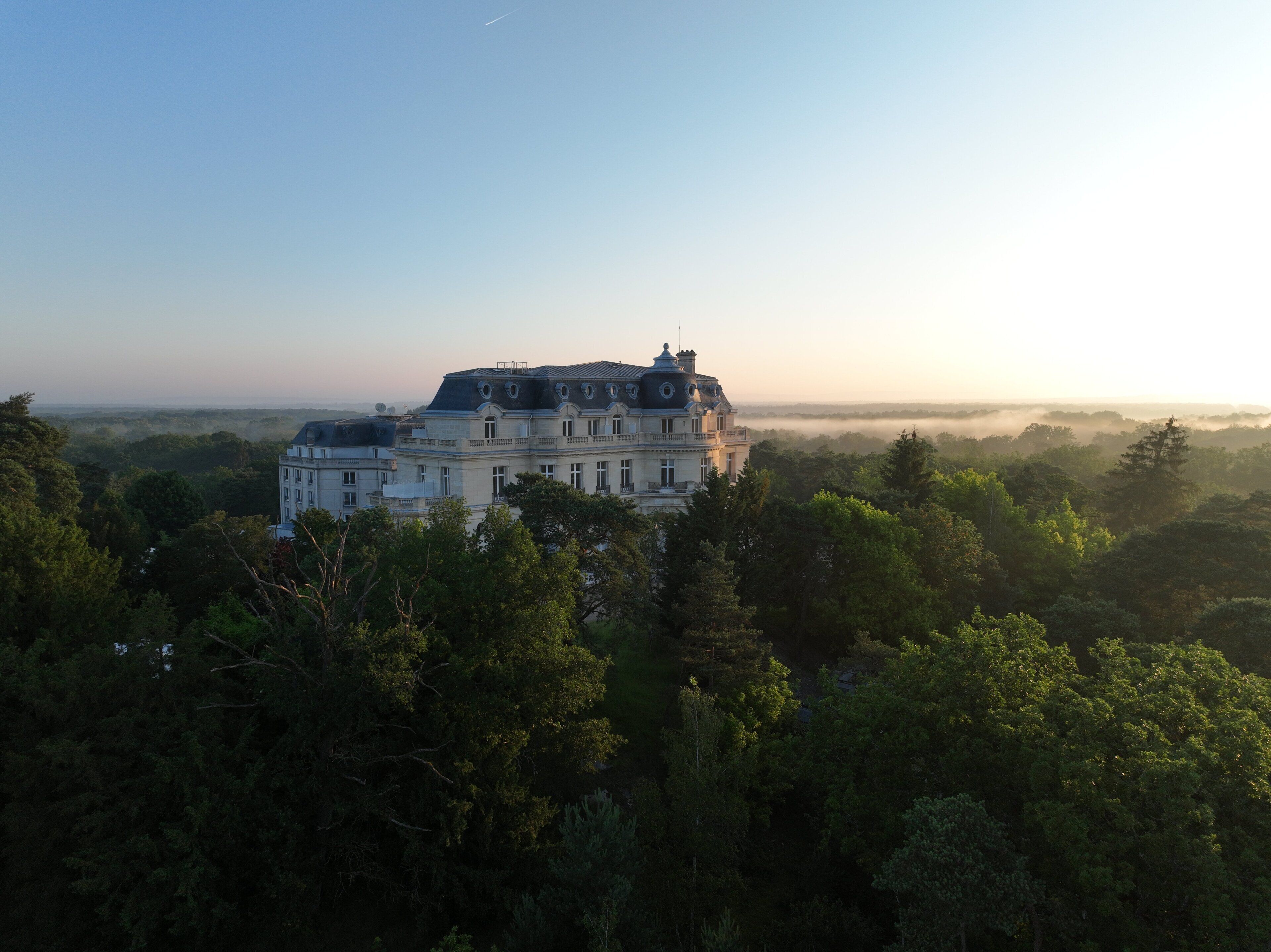 Un grand bâtiment à plusieurs étages, entouré d’une forêt dense, sous un ciel dégagé.