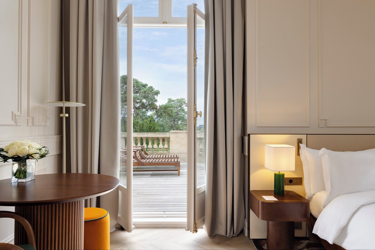 Una habitación de hotel con cama, una mesita de noche con lámpara, una mesa de madera con un florero y vistas a un balcón exterior.