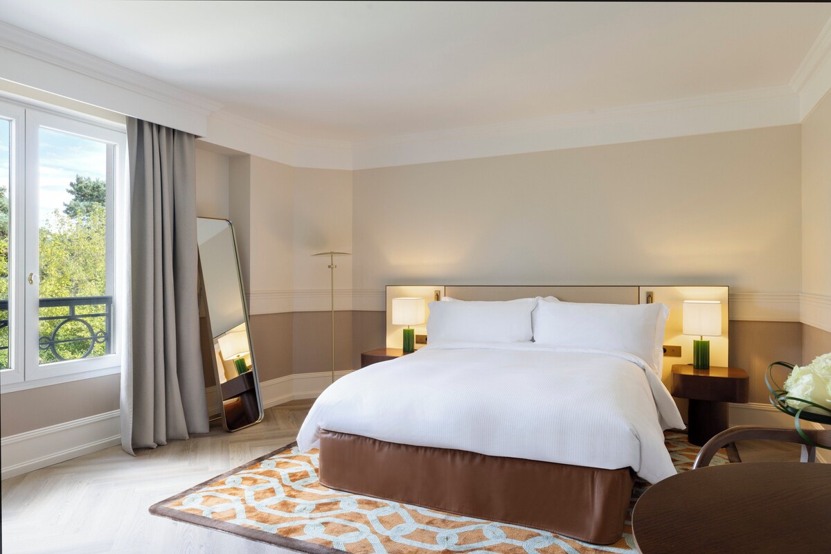 Una habitación de hotel con una cama grande, dos mesitas de noche con lámparas, un espejo, una ventana con cortinas y vistas a zonas verdes.