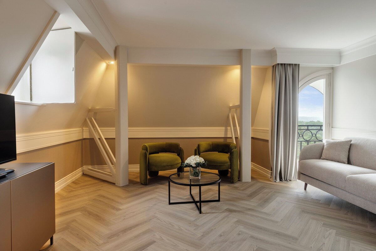 Una habitación de hotel moderna con suelo de madera en patrón espiga, un sofá, dos sillones, una pequeña mesa redonda con un jarrón de flores, un televisor en un mueble bajo y un balcón con cortinas.