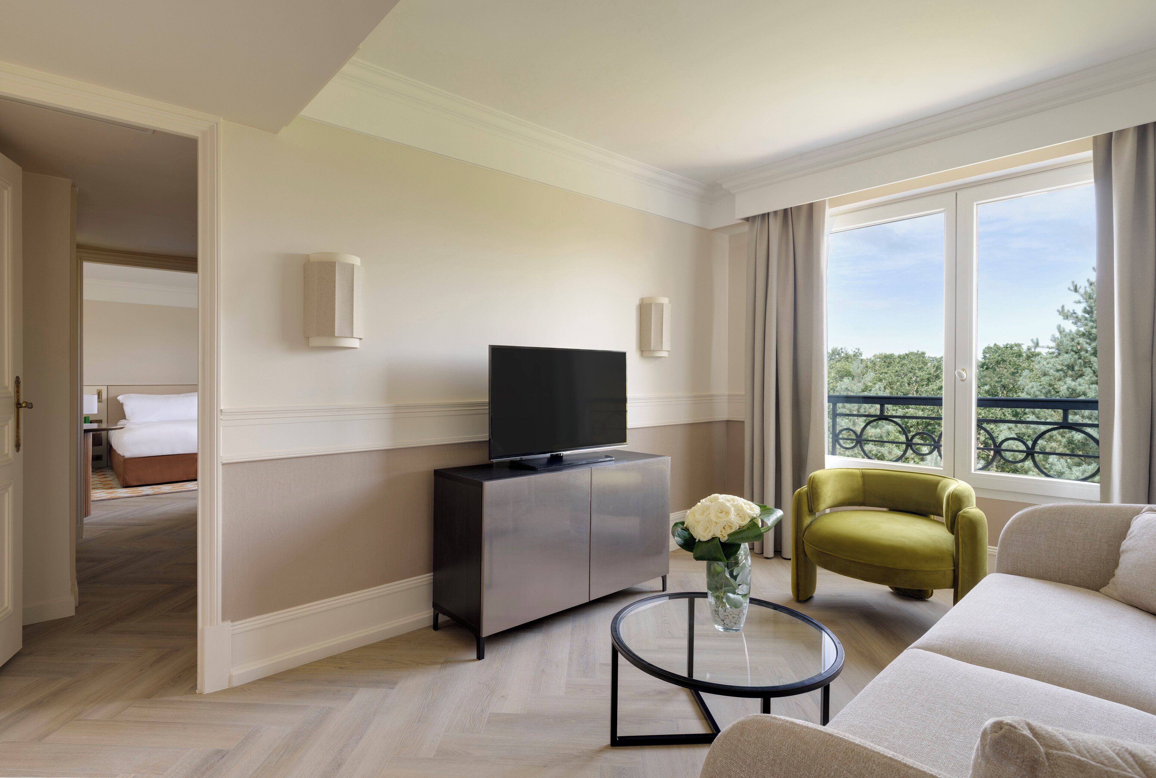 Una habitación de hotel moderna con un sofá, un televisor en un mueble, una pequeña mesa redonda con un florero y un balcón con vistas.